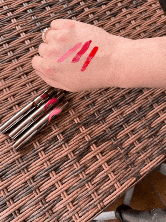 A hand with four different shades of lipstick swatches on the back, placed above three lipstick tubes on a woven surface.