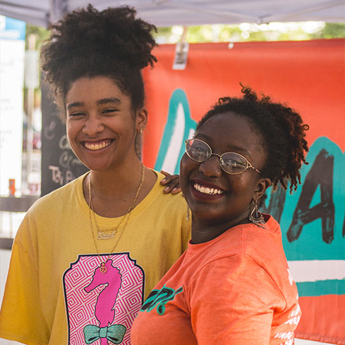 Two women standing and smiling at an outdoor event, one wearing a yellow shirt with a pink design and the other wearing an orange shirt and glasses.