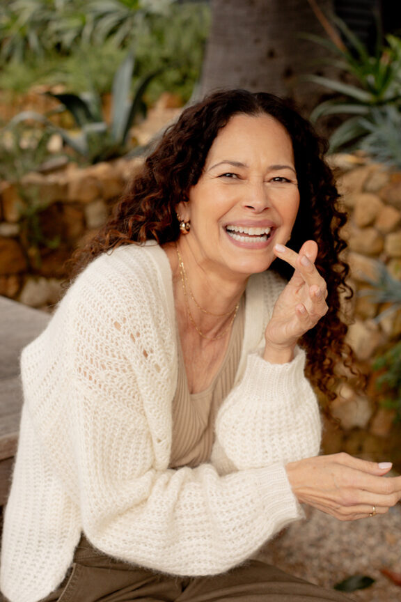 A joyful woman in a white sweater laughing, sitting outdoors with a stone wall in the background.