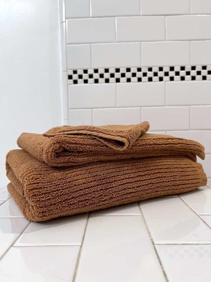 Two folded organic towels are stacked on a white tiled surface in front of a white tile wall with a black and white checkerboard accent.