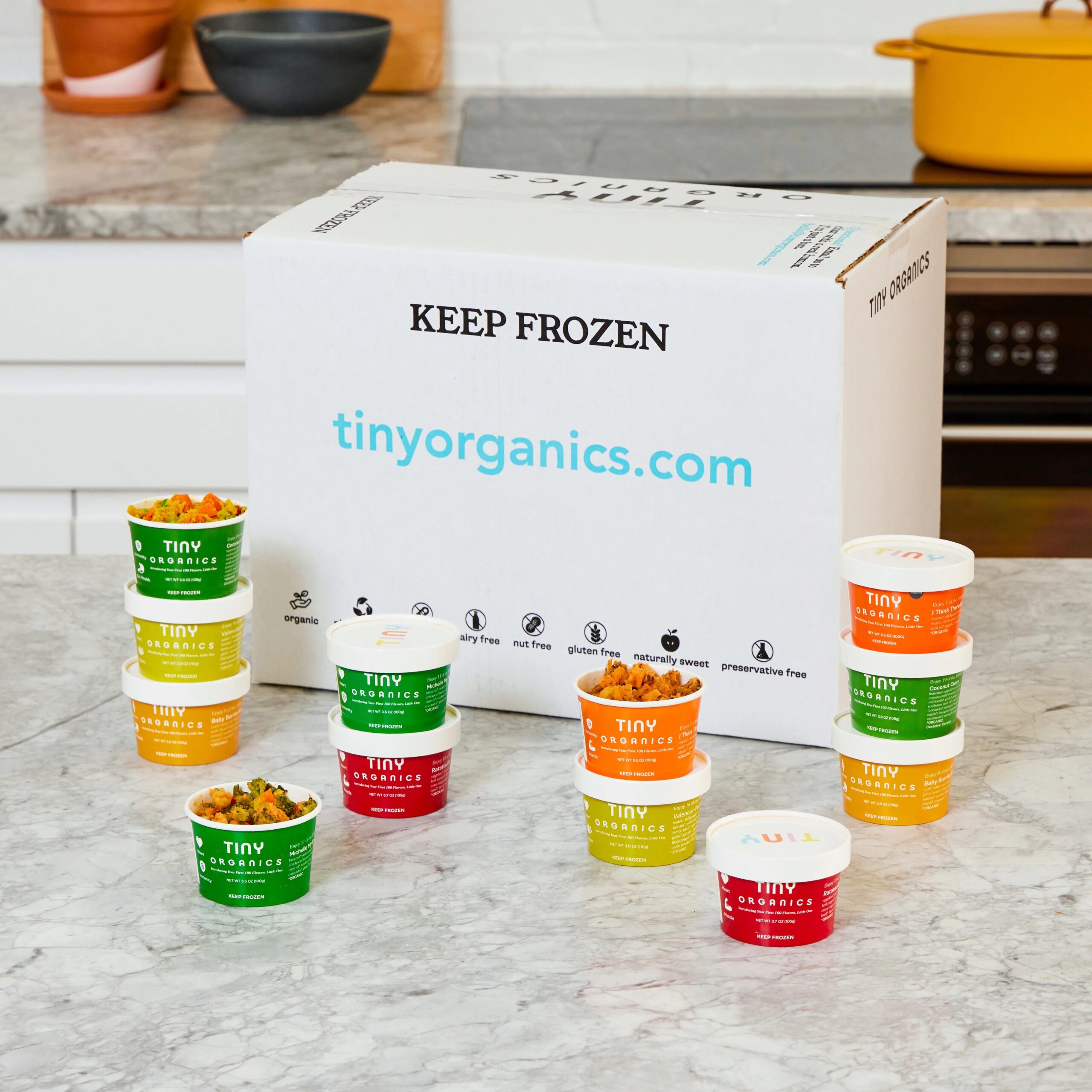 A box of the products sits on a counter surrounded by the containers of food in stacks.
