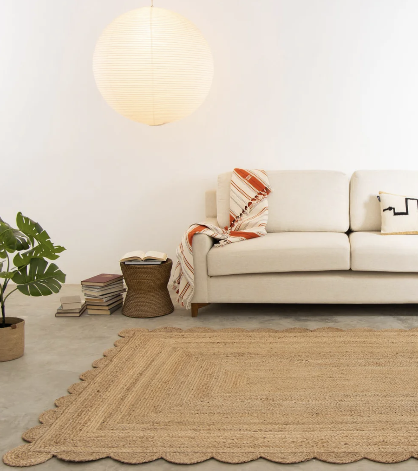 A neutral-toned living room with a beige sofa, patterned throw, potted plant, stack of books, woven basket, and a large round paper lantern on the wall above a scalloped jute rug.