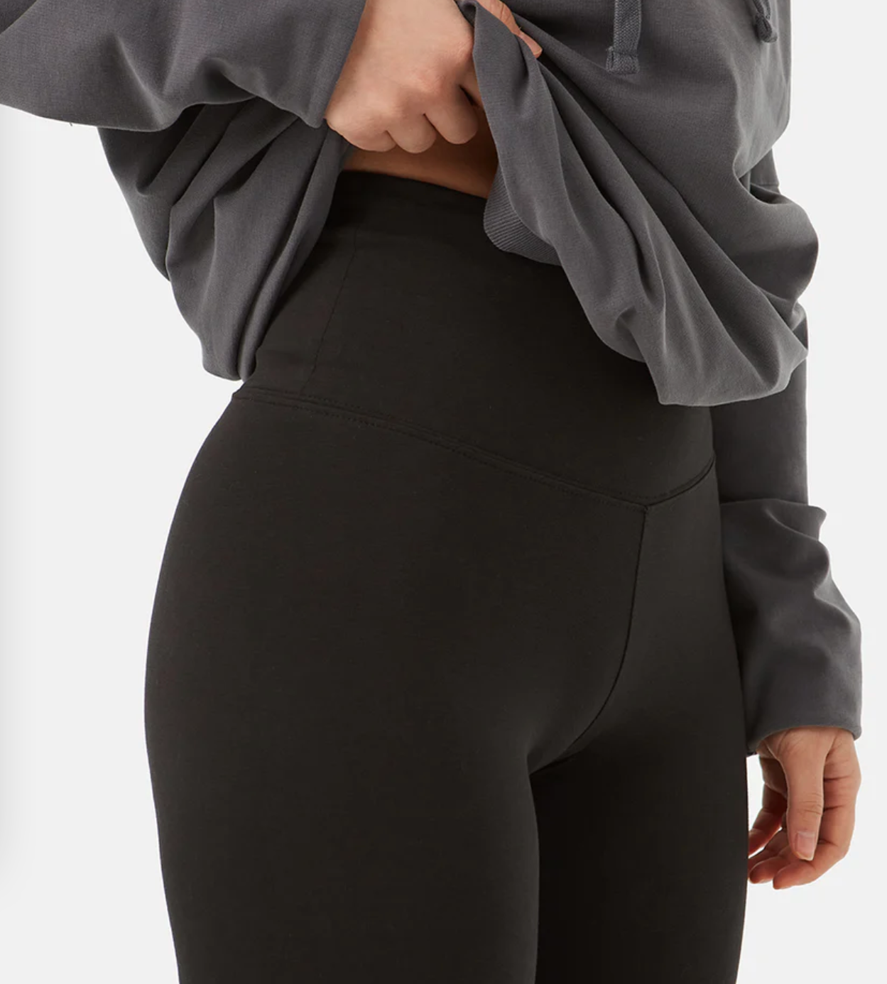 A model lifts her oversized gray hoodie to show the top of her black cotton leggings.