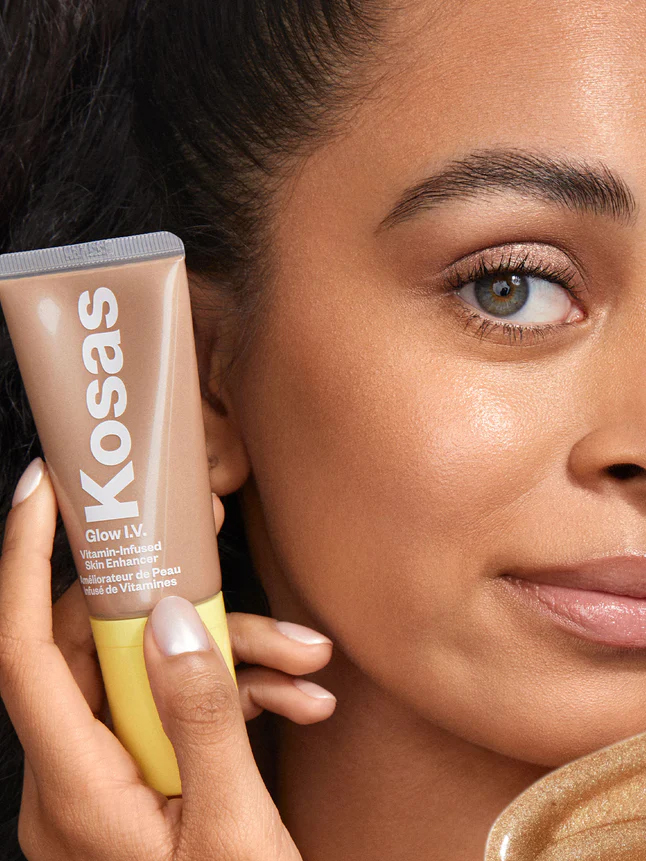 A model holds a tube of Kosas highlighter next to her cheek.