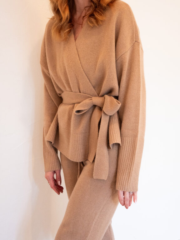 Person wearing a tan knit wrap sweater with a tied waist belt and matching knit pants, standing against a plain light background.