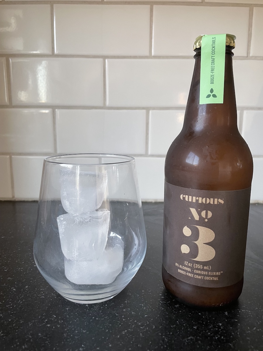 An empty wine tumbler with ice in it, next to a bottle of Curious Elixirs cocktails, set on a kitchen counter.