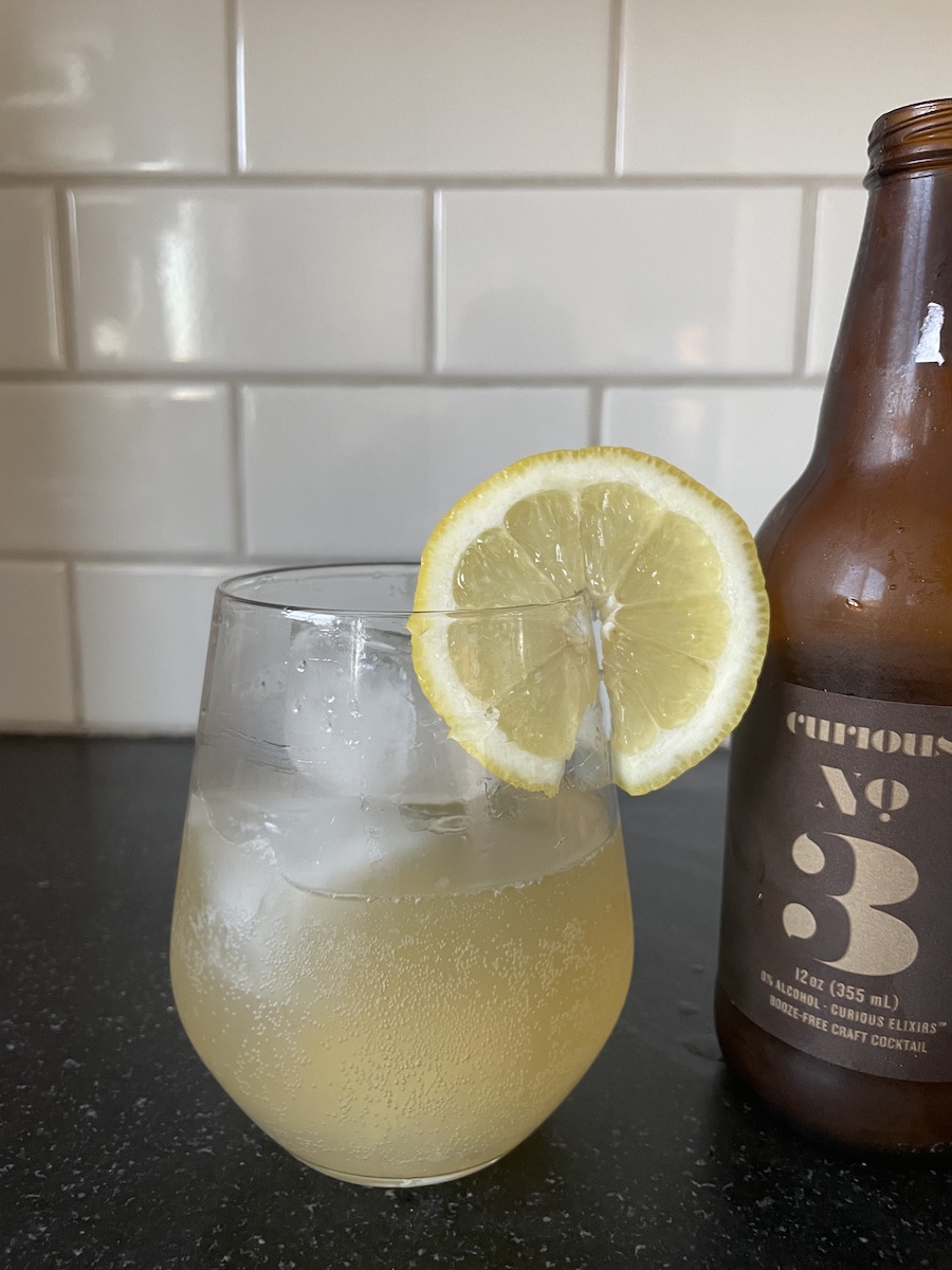 A wine tumbler filled with Curious Elixirs cocktail and garnished with a lemon slice, next to an open bottle of Curious Elixirs on a kitchen counter.