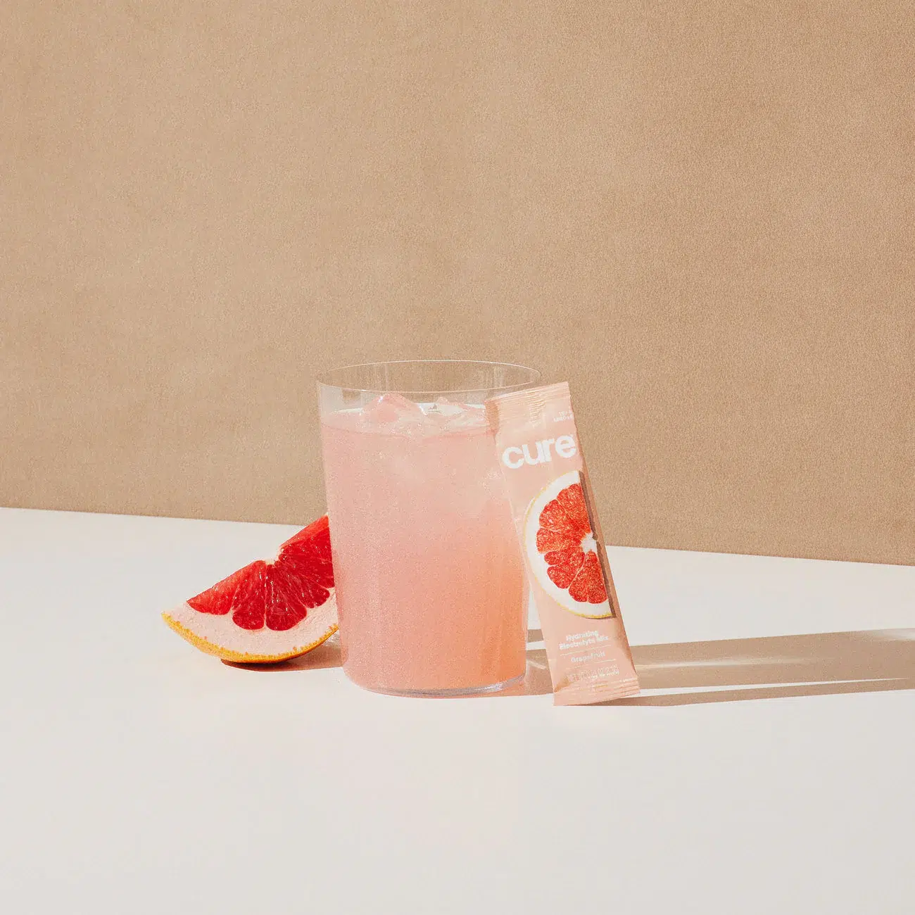 A glass of pink grapefruit drink with ice, a grapefruit slice, and a grapefruit-flavored drink mix packet on a white surface against a beige background.