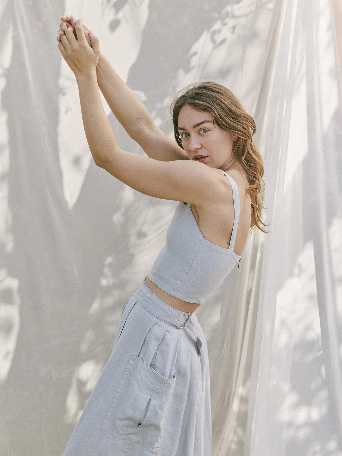 A woman in a light blue outfit stretches her arms upwards, standing among softly draped white curtains.
