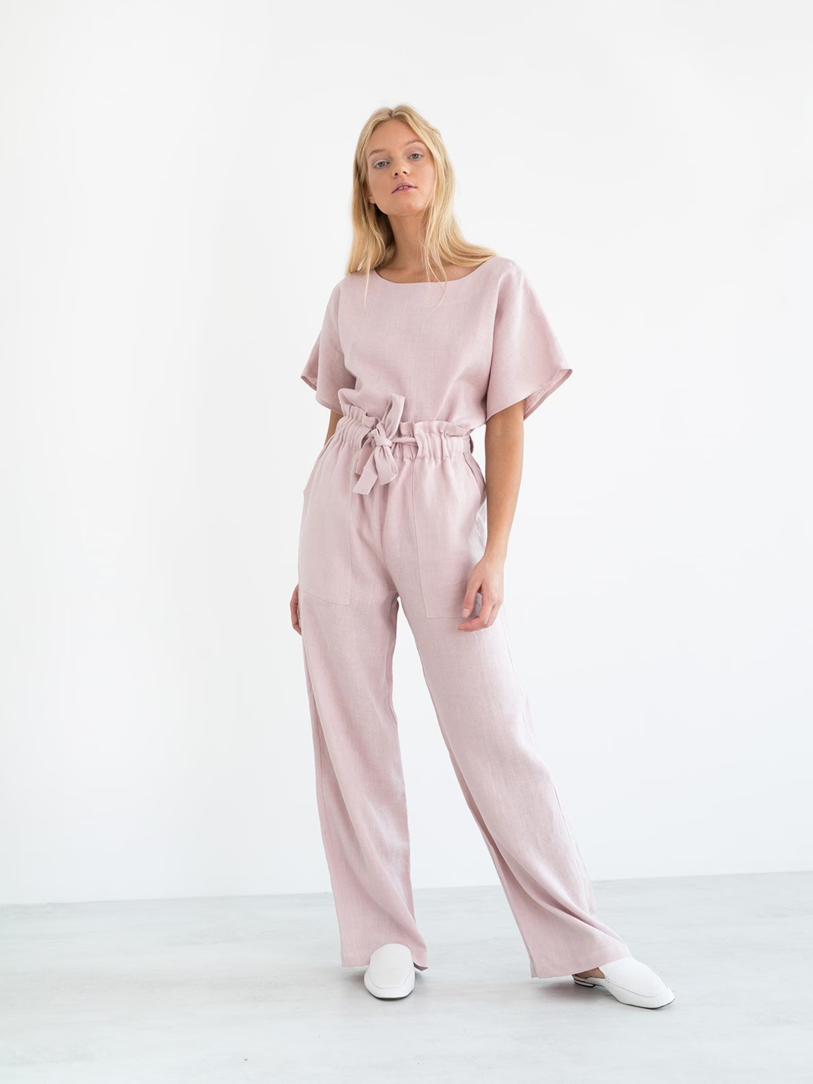 A woman in a light pink jumpsuit stands against a white background, hands slightly apart, wearing white shoes.