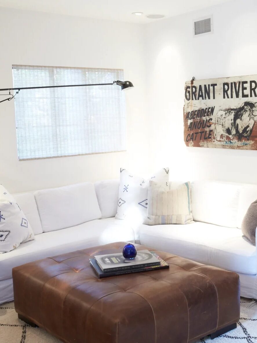 Minimalist living room with a large white sofa, brown leather ottoman, a rustic farm-themed wall sign, and a light window shade.