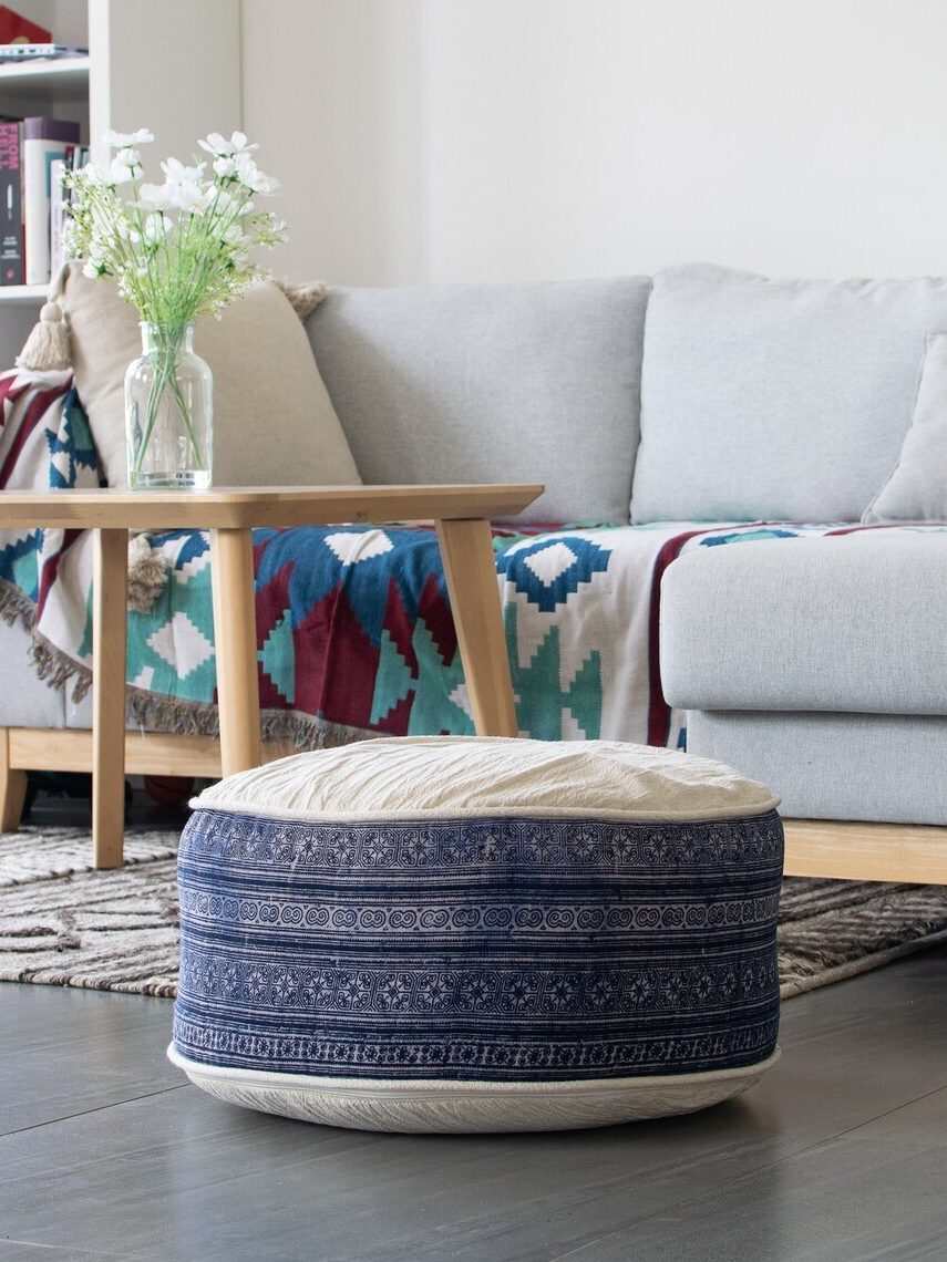 Cozy living room with a gray sofa, patterned pouf and throw blanket, wooden coffee table with flowers, and a bookshelf in the background.