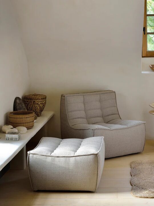 A minimalist living room featuring a gray lounge chair with an ottoman, a wooden side table, a long bench with baskets, and a small window.