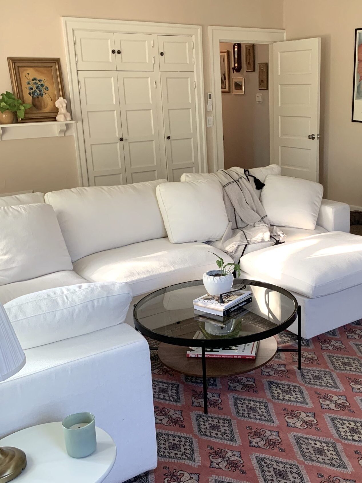 A living room with a white sectional sofa, a glass coffee table, a brown armchair, and a red patterned rug. Artwork and shelves with decor are on the walls. A dog bed is on the floor.