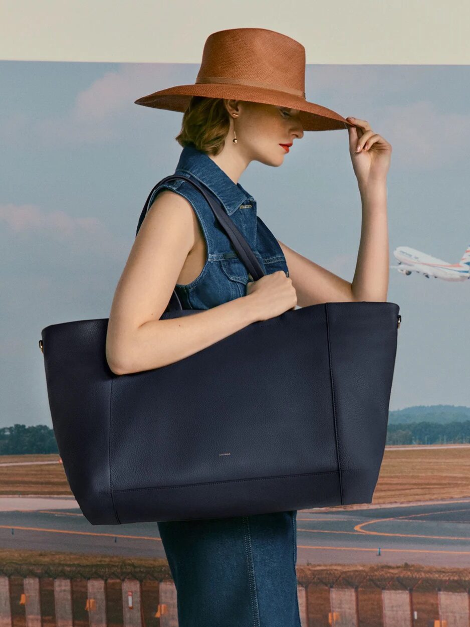 A woman in a wide-brimmed hat and denim vest carries a large tote bag, touching her hat, with an airplane and runway in the background.