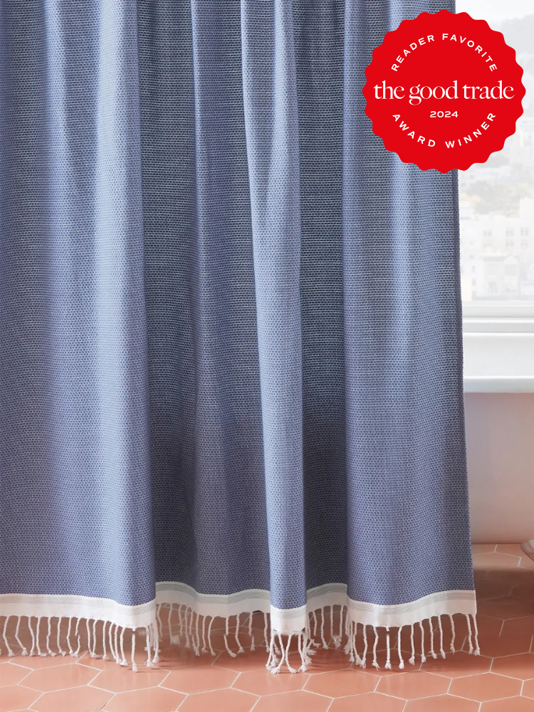 Blue shower curtain with white fringed bottom, hanging in a bathroom with terracotta floor tiles. A red seal in the upper right reads "Reader Favorite, the good trade 2024, Award Winner".