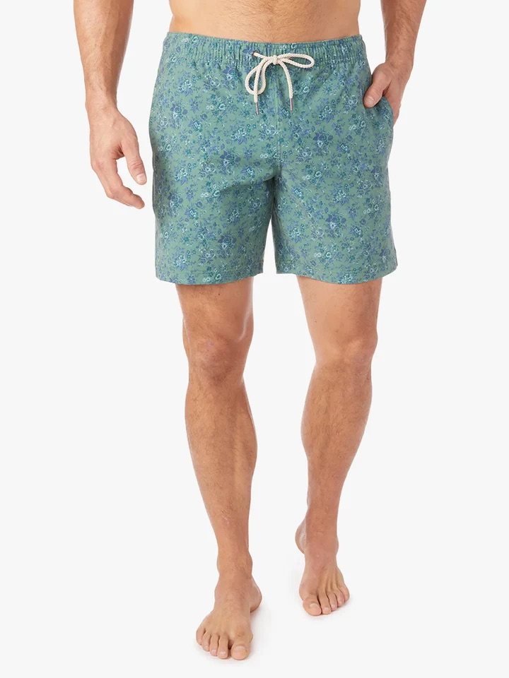 A person is wearing green floral swim trunks with a drawstring, standing against a white background.