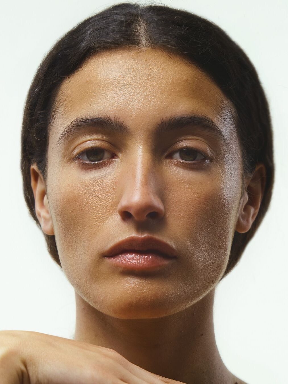 A close-up of a person with a neutral expression, dark hair, and clear skin. The background is plain and light-colored.