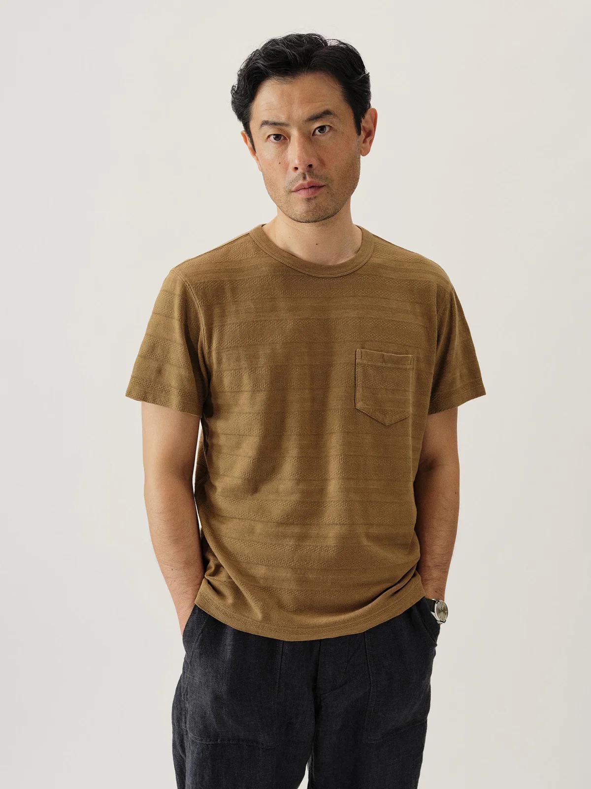 Man standing against a plain background, wearing a brown short-sleeve shirt with a pocket and dark pants, with his hands in his pockets.
