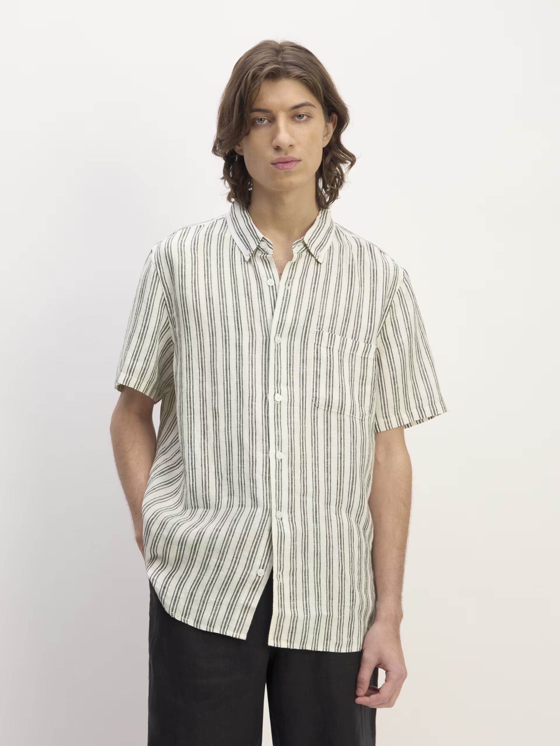 A person with long hair wears a short-sleeve, button-up shirt with vertical black and white stripes, paired with dark pants, standing against a plain white background.