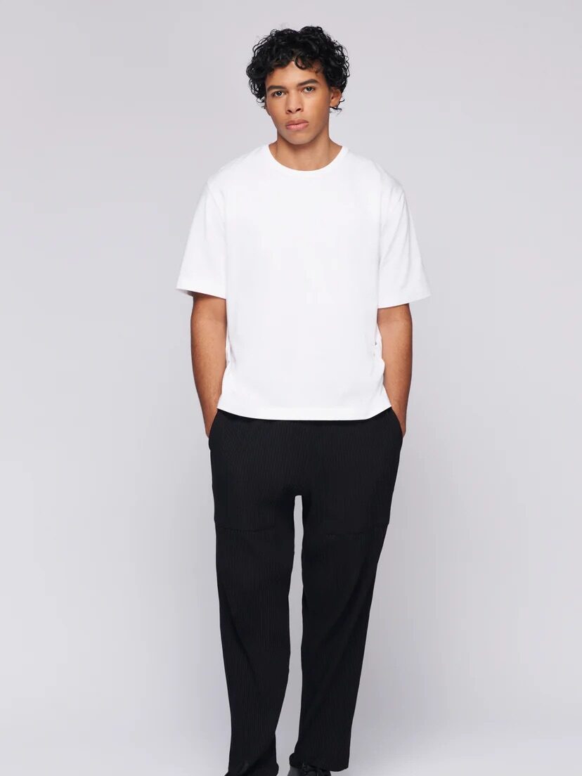 A person stands against a plain background, wearing a white short-sleeved t-shirt, black pants, and black shoes. The person has curly dark hair and appears to be looking straight ahead.