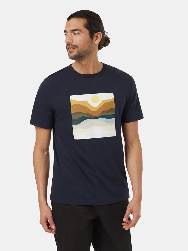 A man with long hair tied back is wearing a navy blue T-shirt with a graphic of abstract mountains and a sun. He is looking to his left and standing against a plain white background.