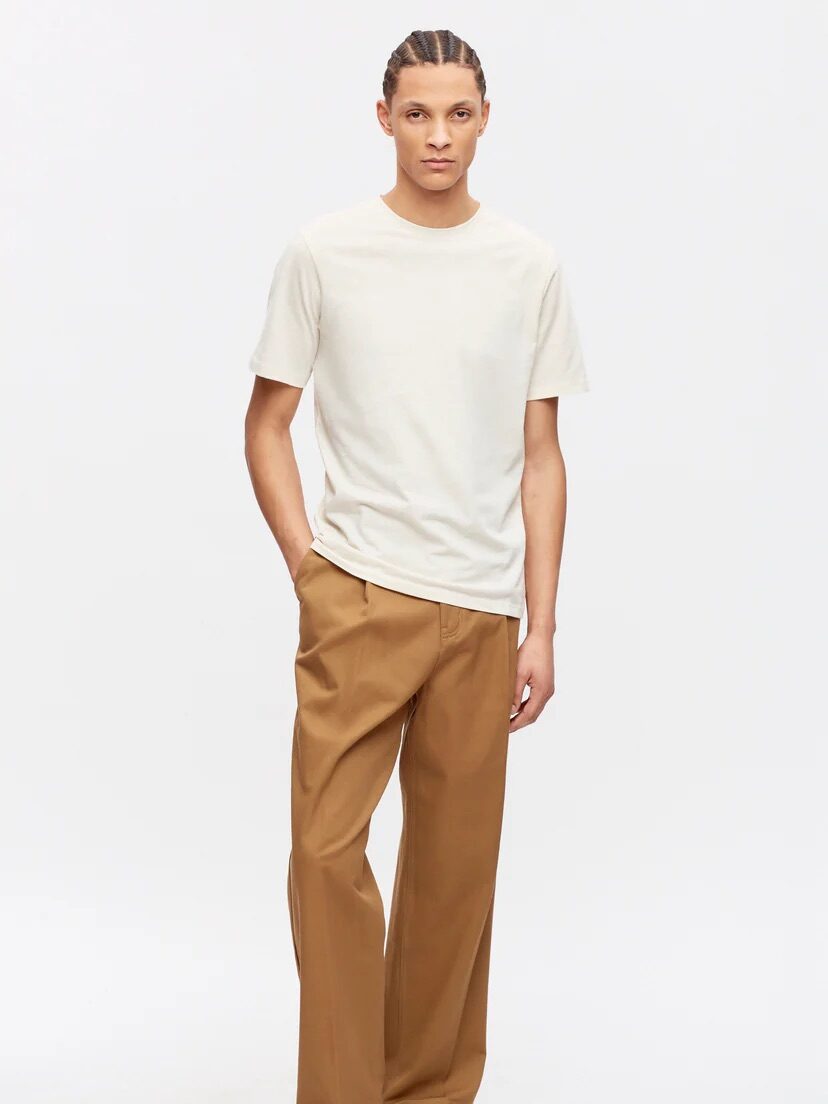 A person with braided hair stands against a plain background, wearing a light beige t-shirt, brown trousers, and black shoes. Their hands are in the pockets of their trousers.