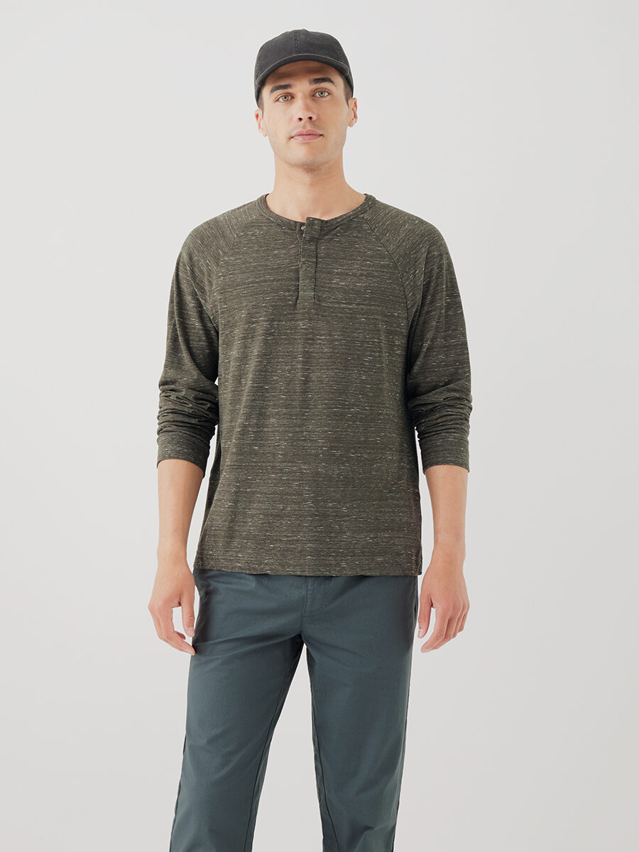 A man wearing a dark green long-sleeve henley shirt with gray pants and a black baseball cap stands against a plain white background.