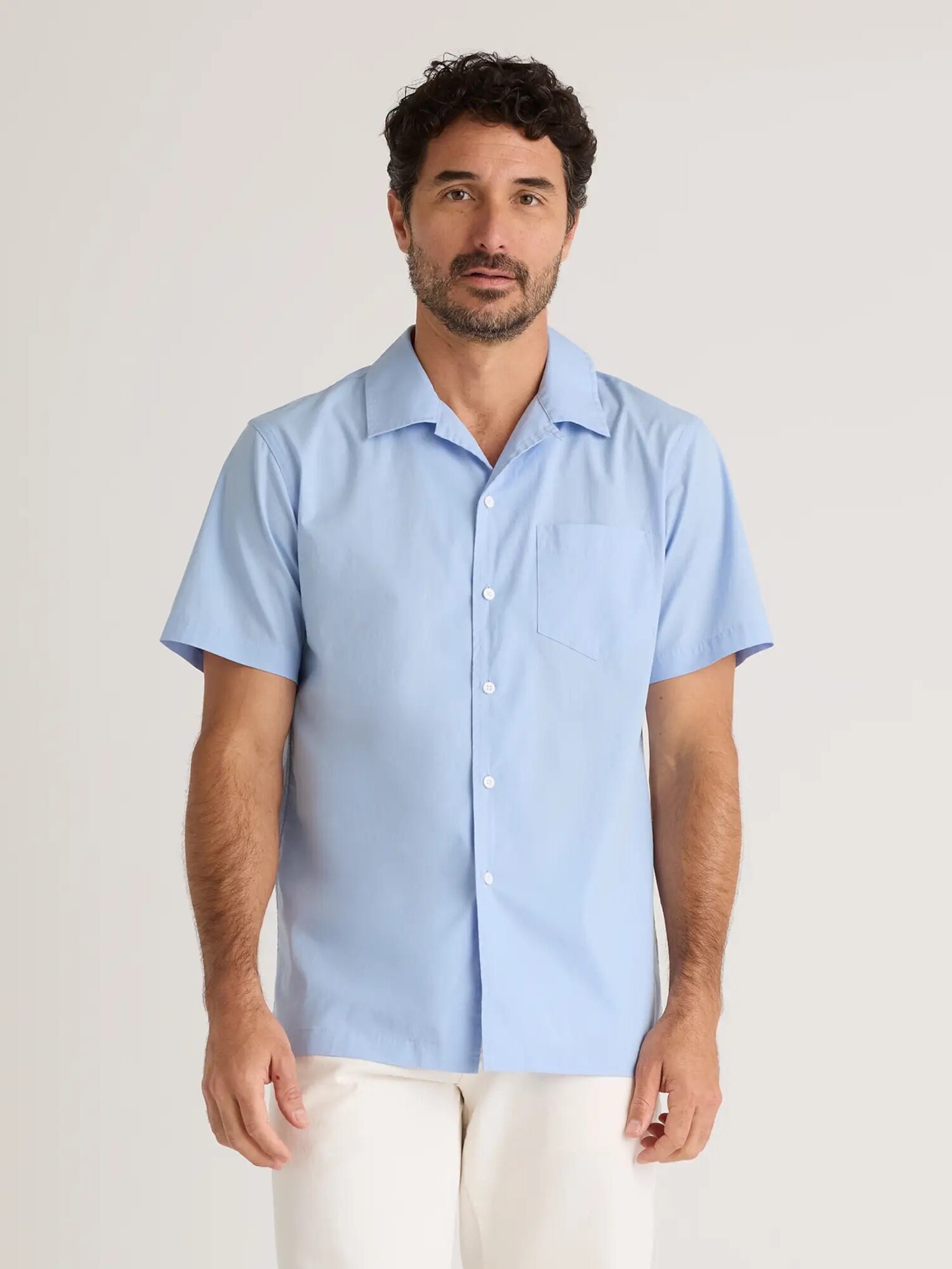 A man with short dark hair and a beard is wearing a light blue, short-sleeve button-up shirt. He is standing against a plain light background.