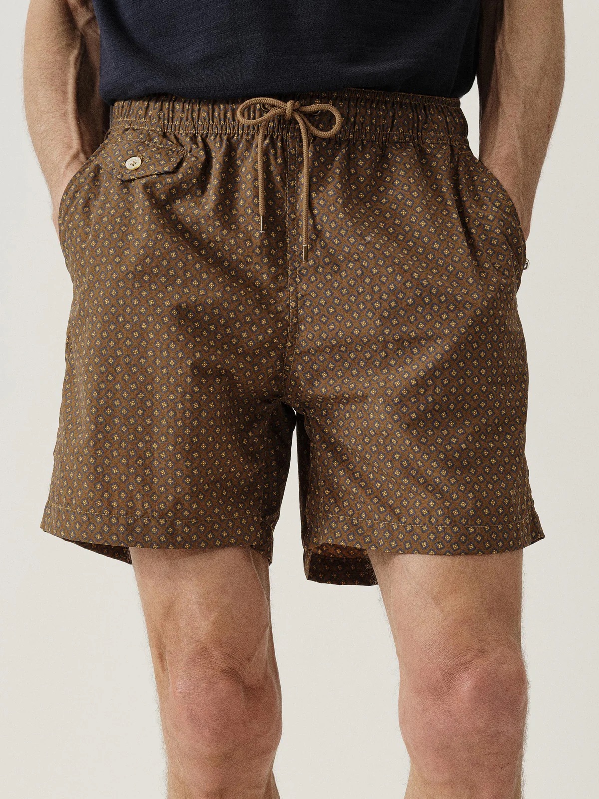 A person wearing brown patterned shorts with hands in pockets. The shorts have a drawstring and a small buttoned pocket on the left side. The person is standing against a plain background.