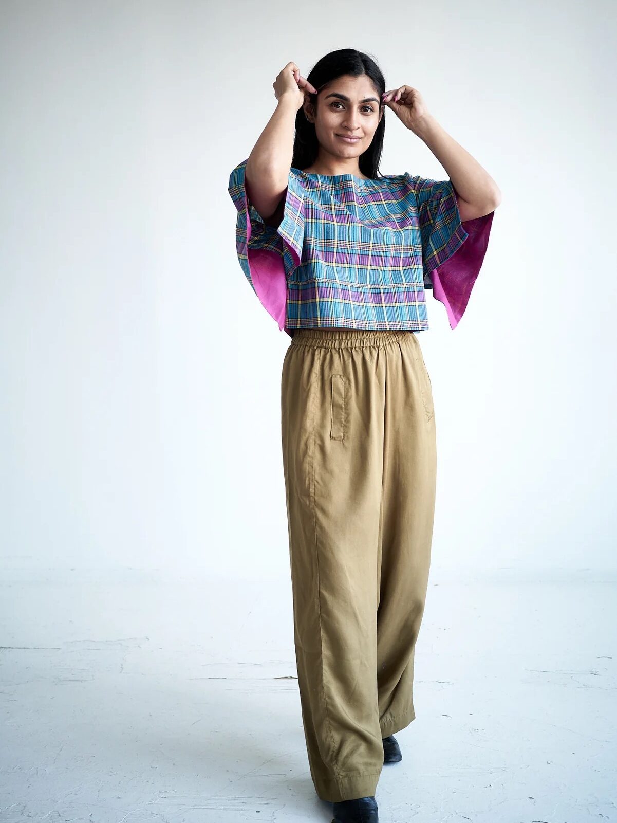 A person stands in a well-lit room adjusting their hair, wearing a multicolored plaid top with wide sleeves and loose-fitting beige pants.