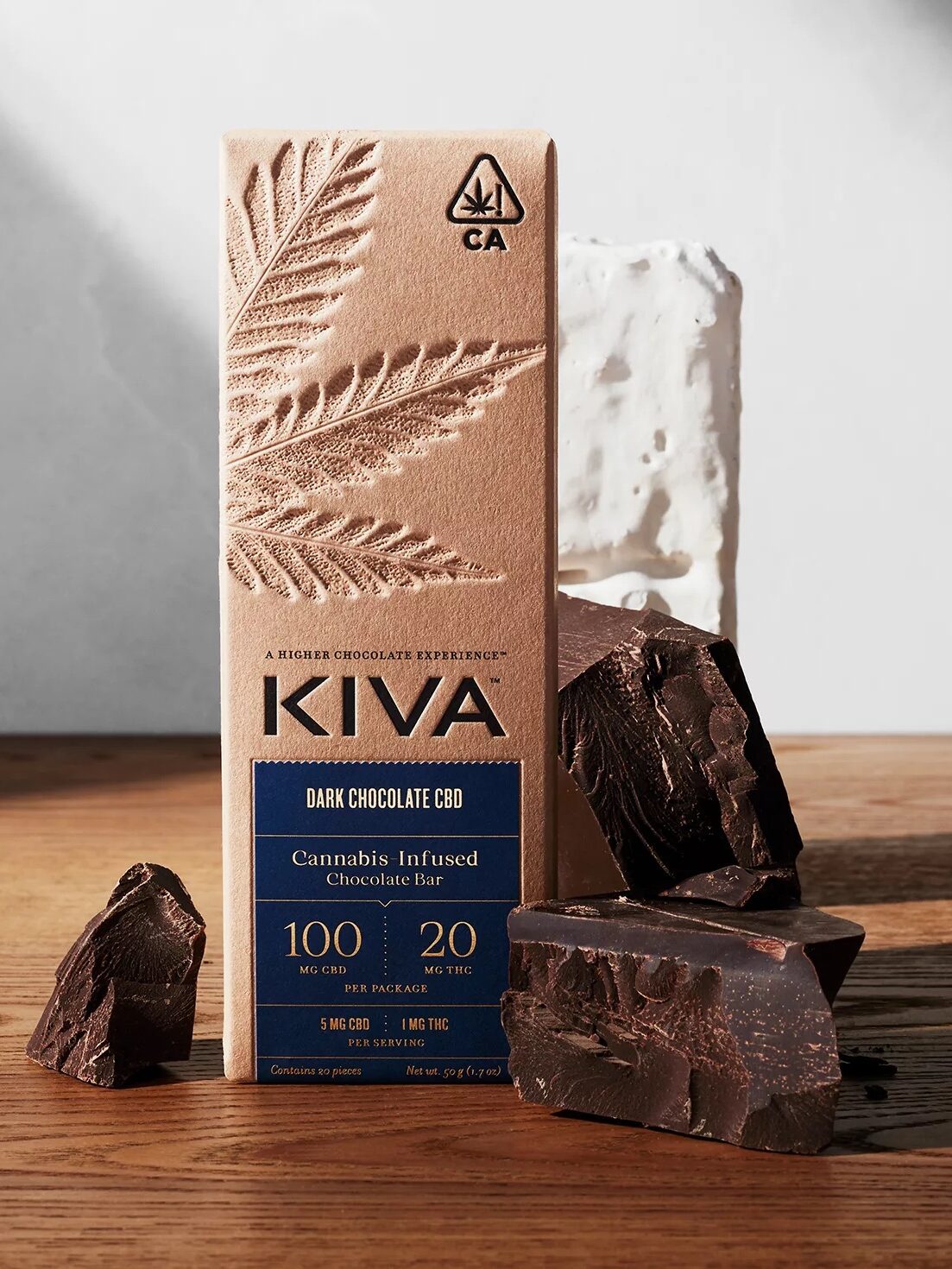A Kiva dark chocolate CBD bar with packaging showing cannabis leaf and dosage details, accompanied by broken chocolate pieces on a wooden surface.