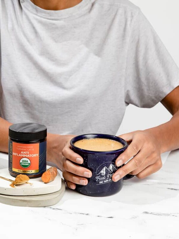 Person in a grey shirt holding a blue mug, with a bowl of cereal, raspberries, a jar of powdered supplement, and a spoon with powder on a white marble table.