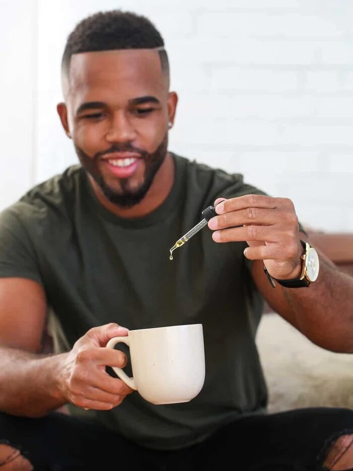 A person with a trimmed beard and green shirt sits on a couch, holding a white mug in one hand and using a dropper to add liquid to it with the other hand.