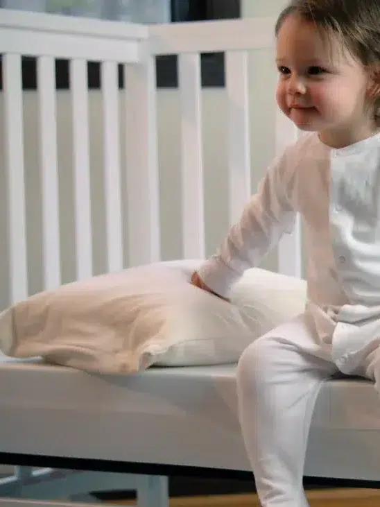 A toddler in white pajamas sits on the edge of a white crib mattress, with a pillow next to them and a crib railing in the background.