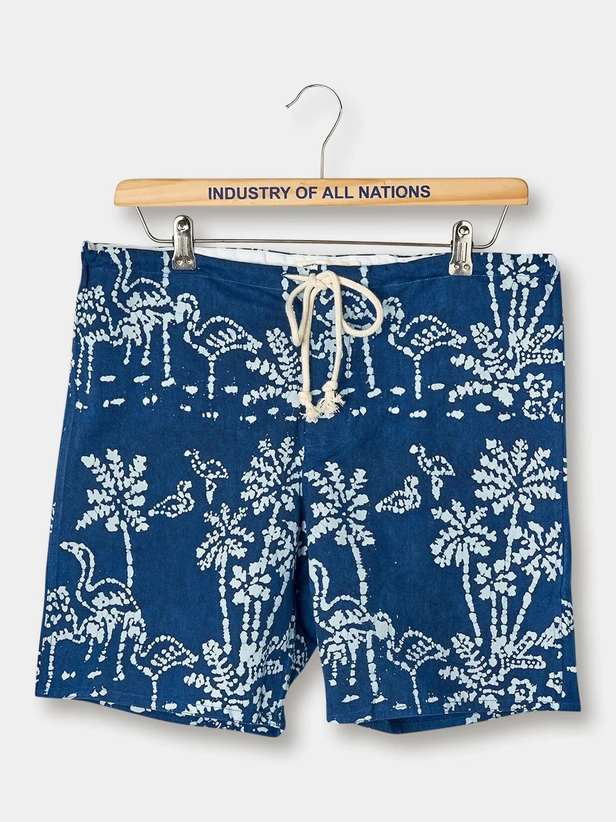 Blue shorts with a white tropical print featuring plants and dinosaurs, hanging from a wooden hanger labeled "Industry of All Nations.
