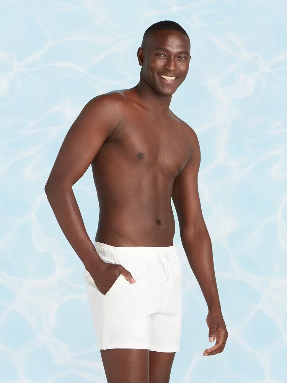 A man in white swim shorts stands smiling against a light blue, water-patterned background.