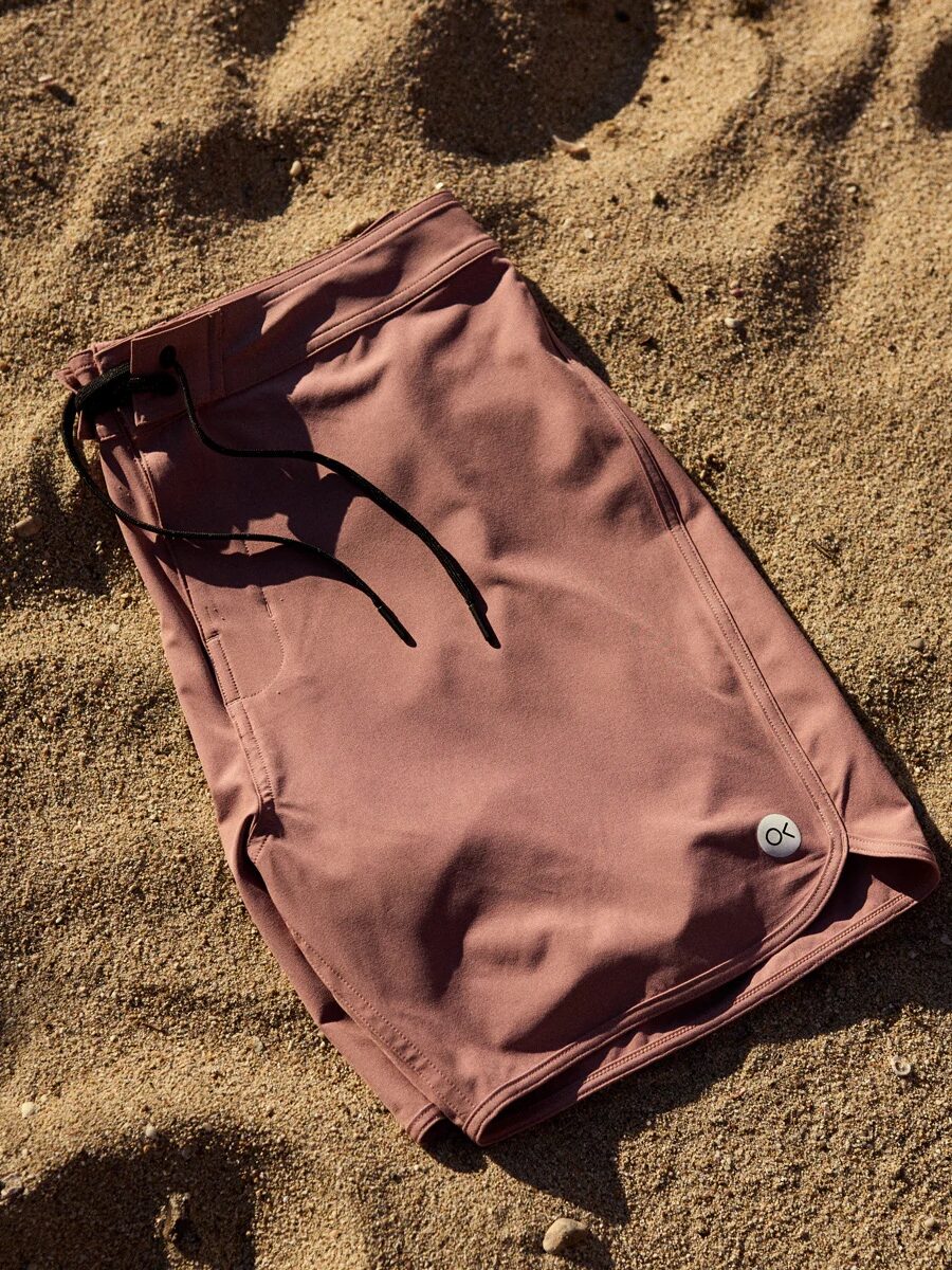 Maroon swim trunks with black drawstrings lie on sandy ground in sunlight.