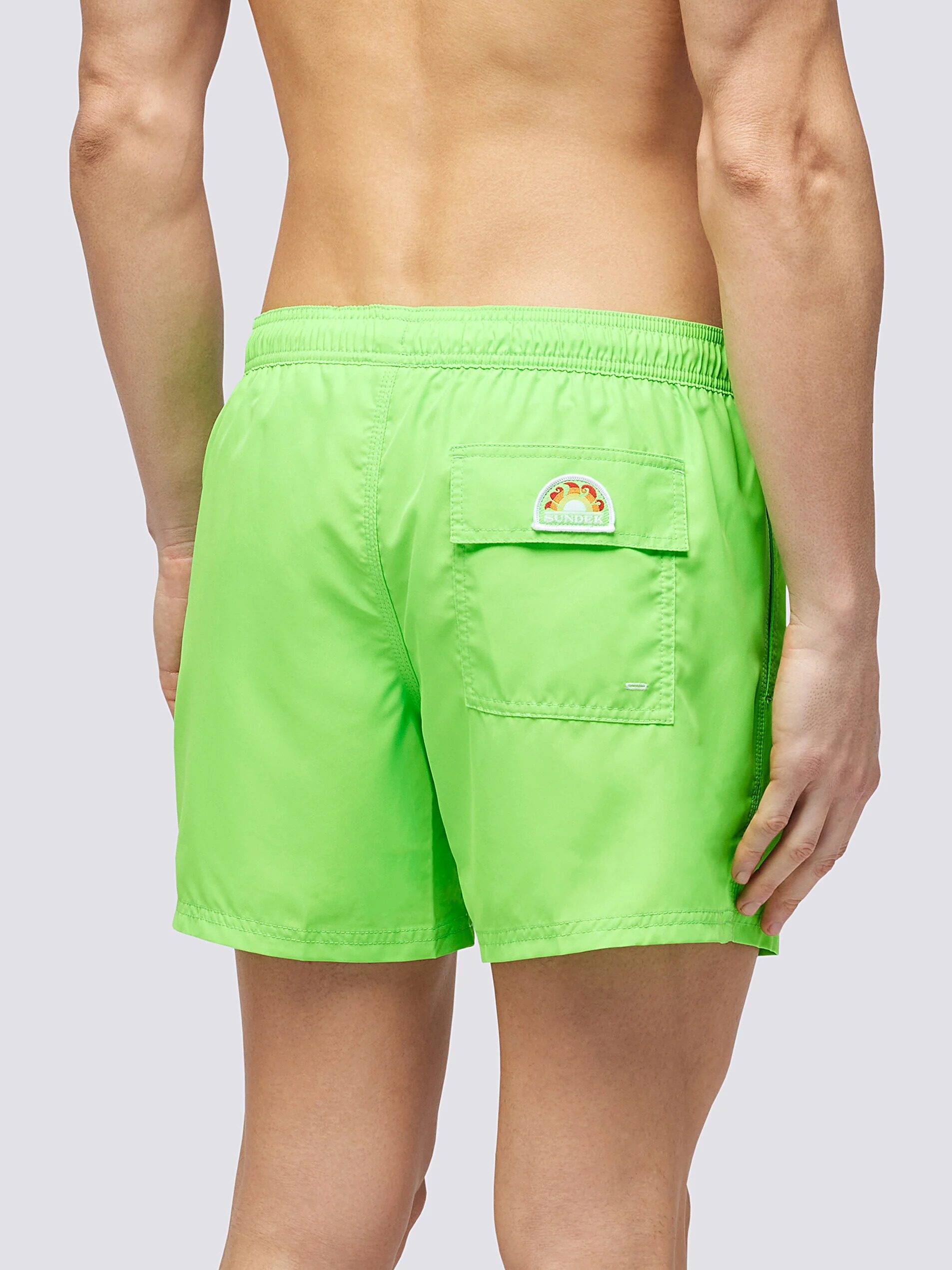 A person wearing bright green swim trunks is shown from the back. The swim trunks have a pocket on the right side with a small embroidered design above it. The waistband is elastic.