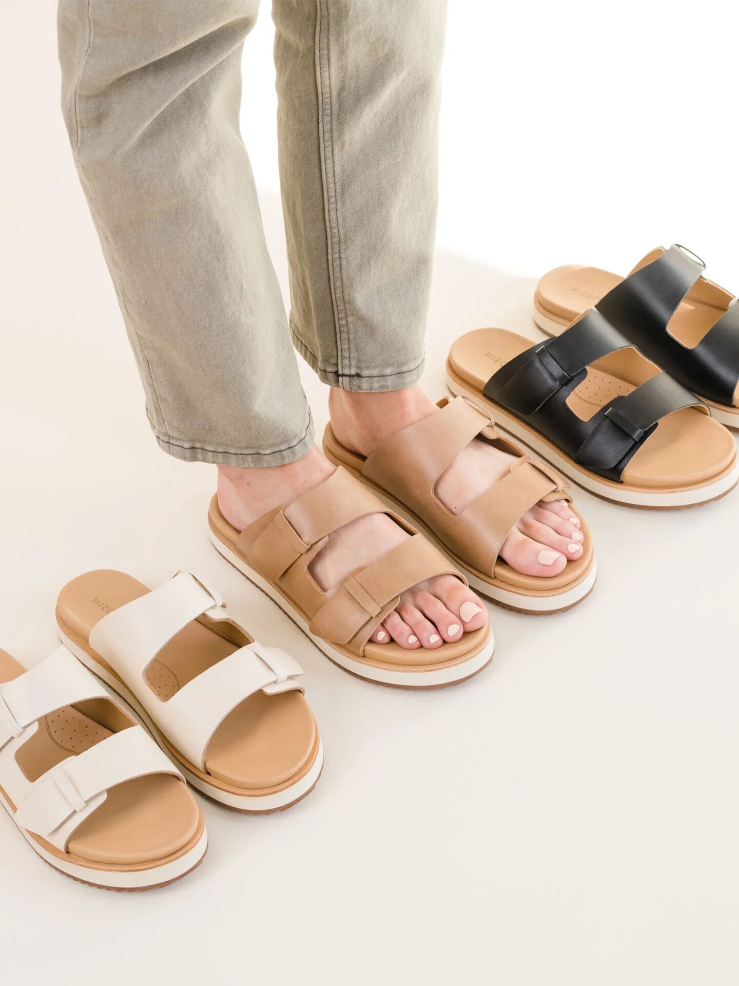 Person wearing beige sandals stands next to additional pairs in black, cream, and another beige. They are dressed in light tan pants on a light surface.