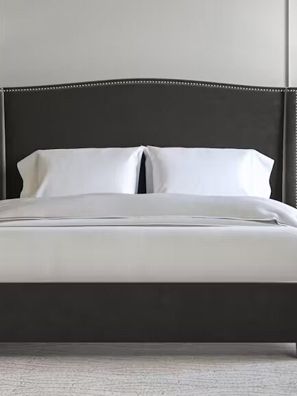 A neatly made bed with white linens, flanked by two bedside tables with identical lamps. The room features a grey upholstered headboard with nailhead trim and a beige armchair on the right.