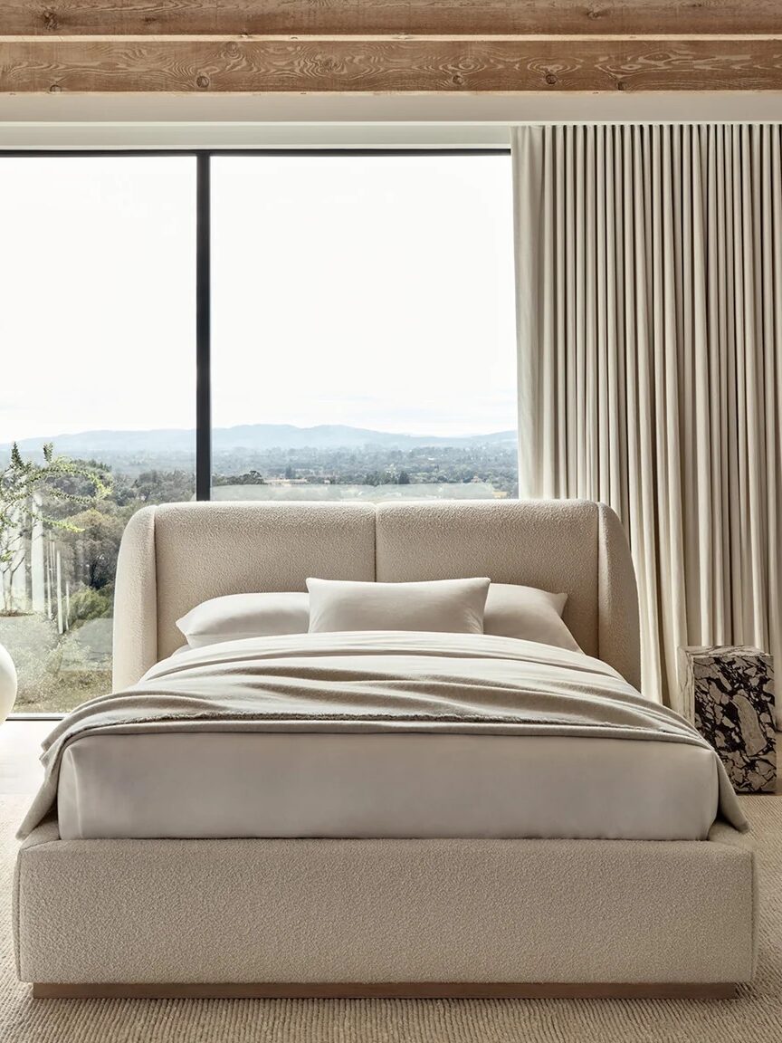 A modern bedroom with a beige bed, large windows, light curtains, a potted plant, and a scenic outdoor view.