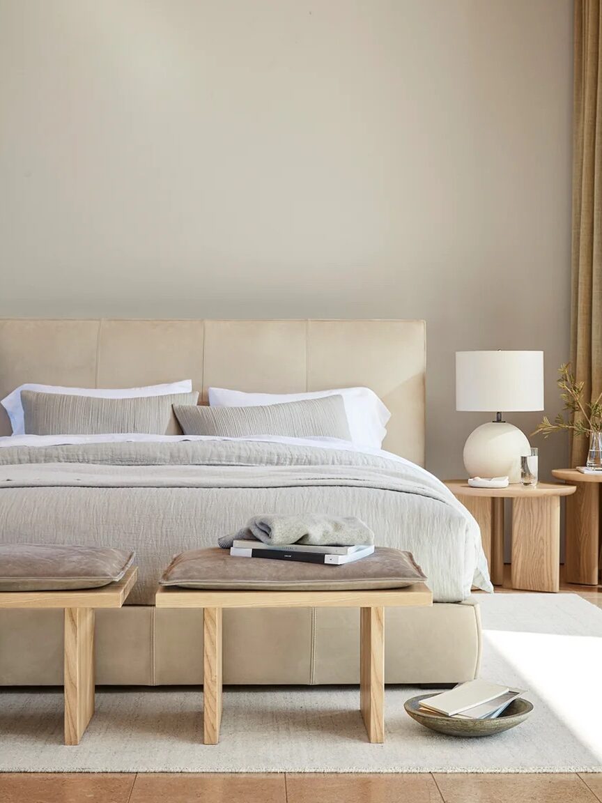 A neatly made bed with beige bedding is in front of a light-colored wall. Two wooden benches are at the foot of the bed, and a wooden nightstand with a lamp and decor is to the right.