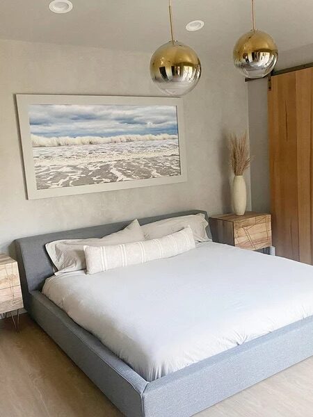 A modern bedroom features a bed with light bedding, wooden nightstands, pendant lights, and a sliding barn door leading to an adjoining bathroom with a glass shower.