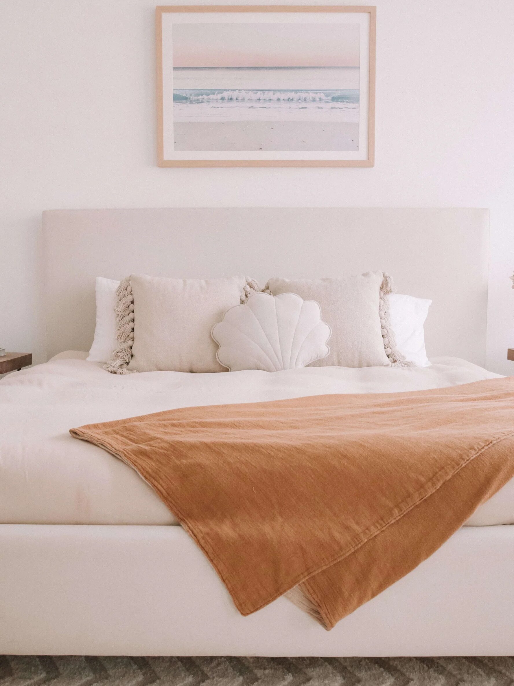 Minimalist bedroom with a white bed, a brown throw blanket, and a seashell pillow. Two side tables hold books and dried flowers. A beach photo hangs on the wall above the bed.