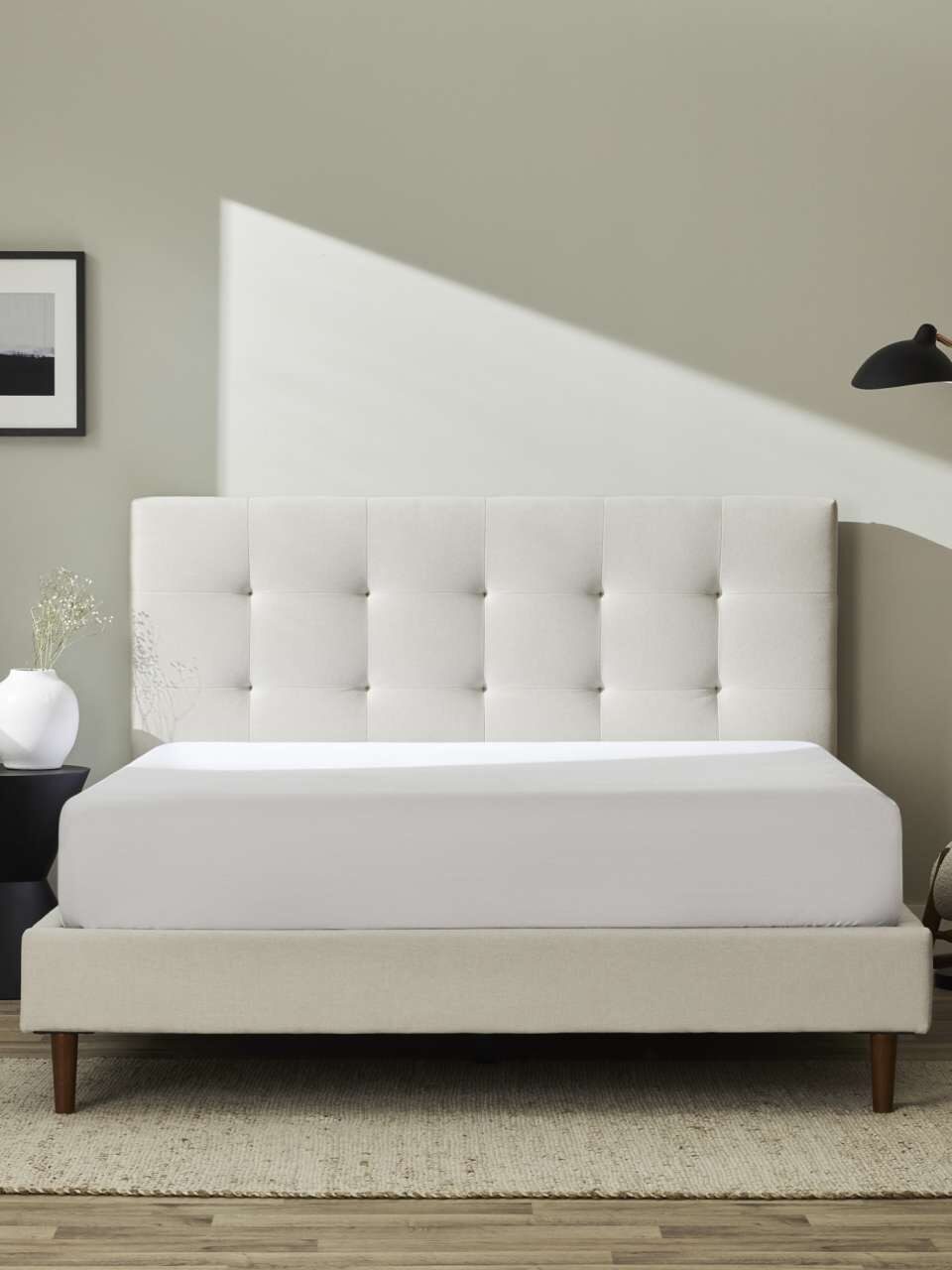 Minimalist bedroom with a beige tufted headboard, white bed, black side table with a vase, modern black floor lamp, and light-colored armchair on a light wood floor with a neutral rug.