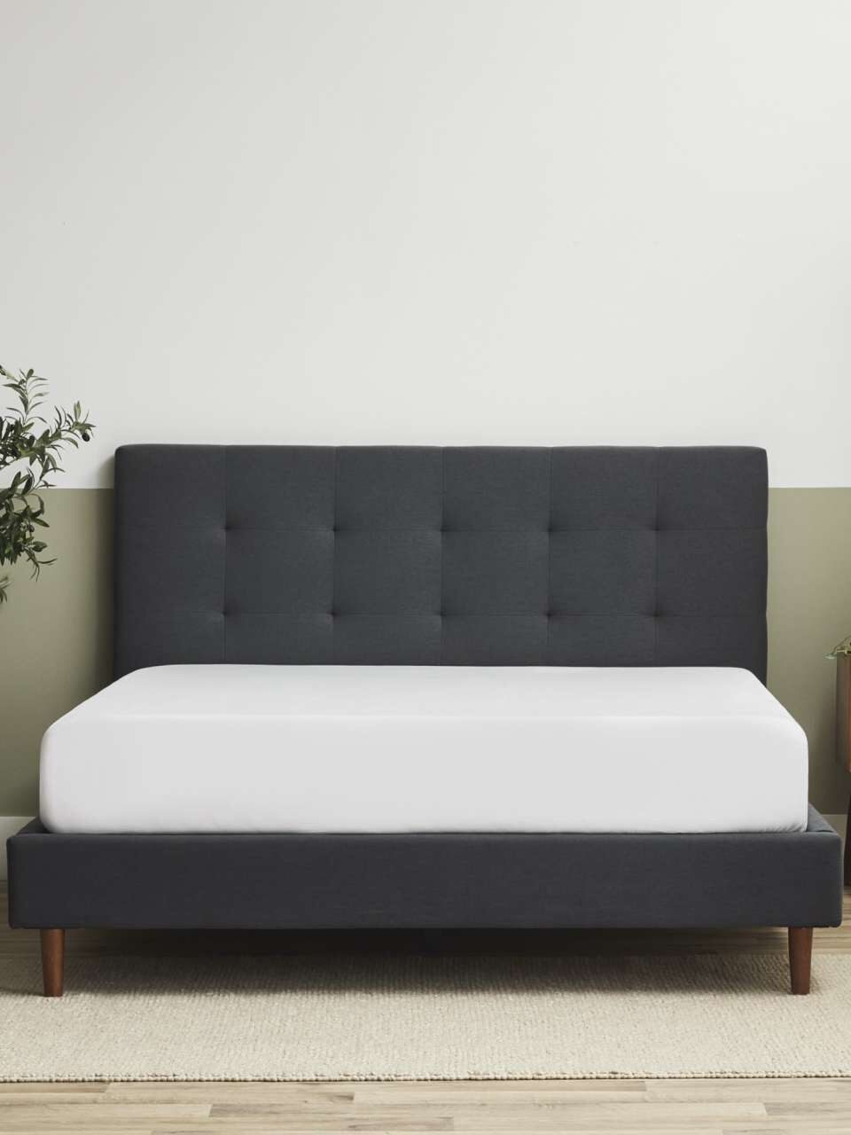 A bedroom with a dark gray upholstered bed, white bedding, a wooden nightstand, a black hanging light, and a small plant in the corner. A painting is on the wall, and a rug covers the wooden floor.