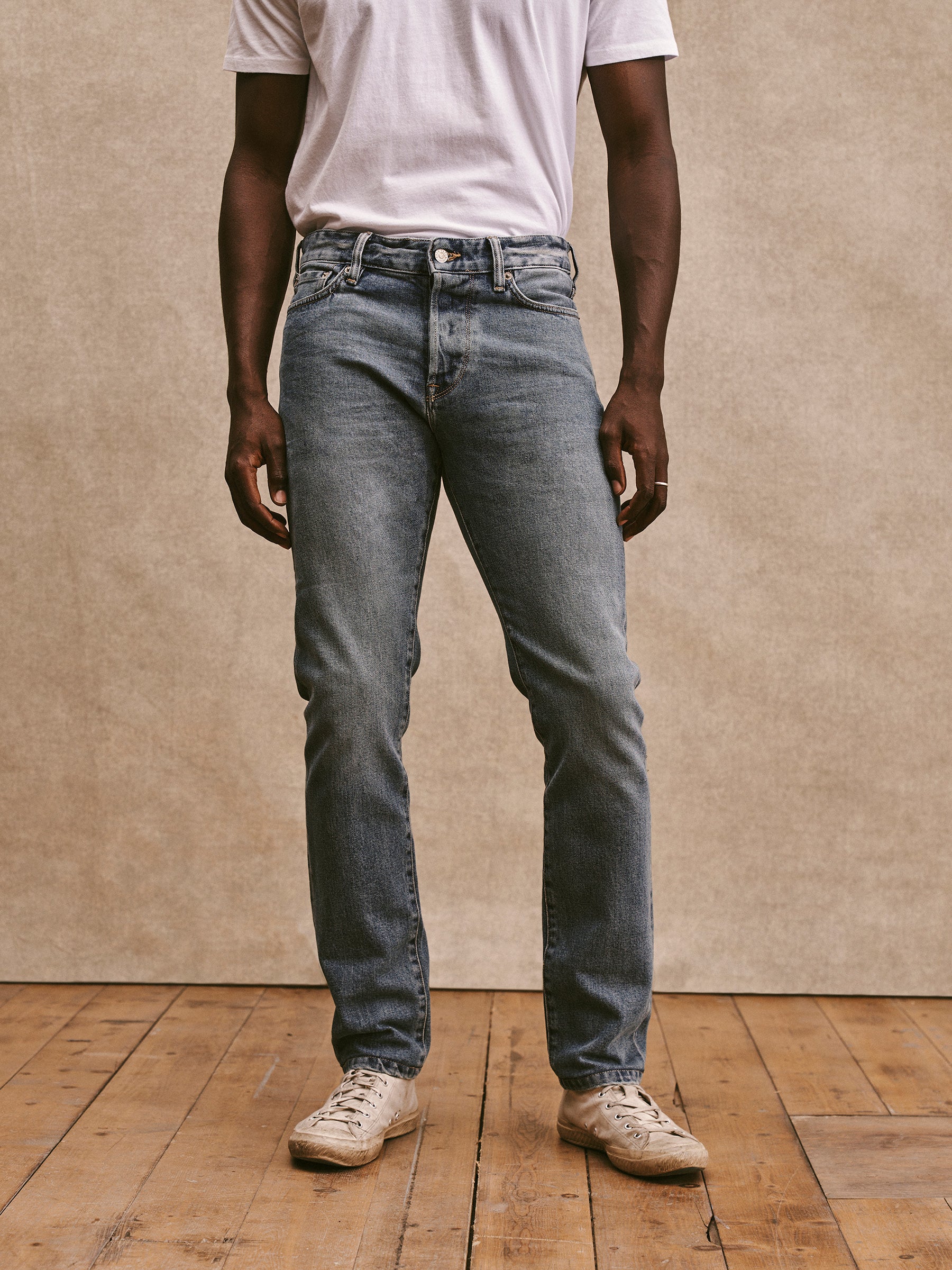 A person wearing a white t-shirt, light blue jeans, and white sneakers stands on a wooden floor with a plain backdrop.