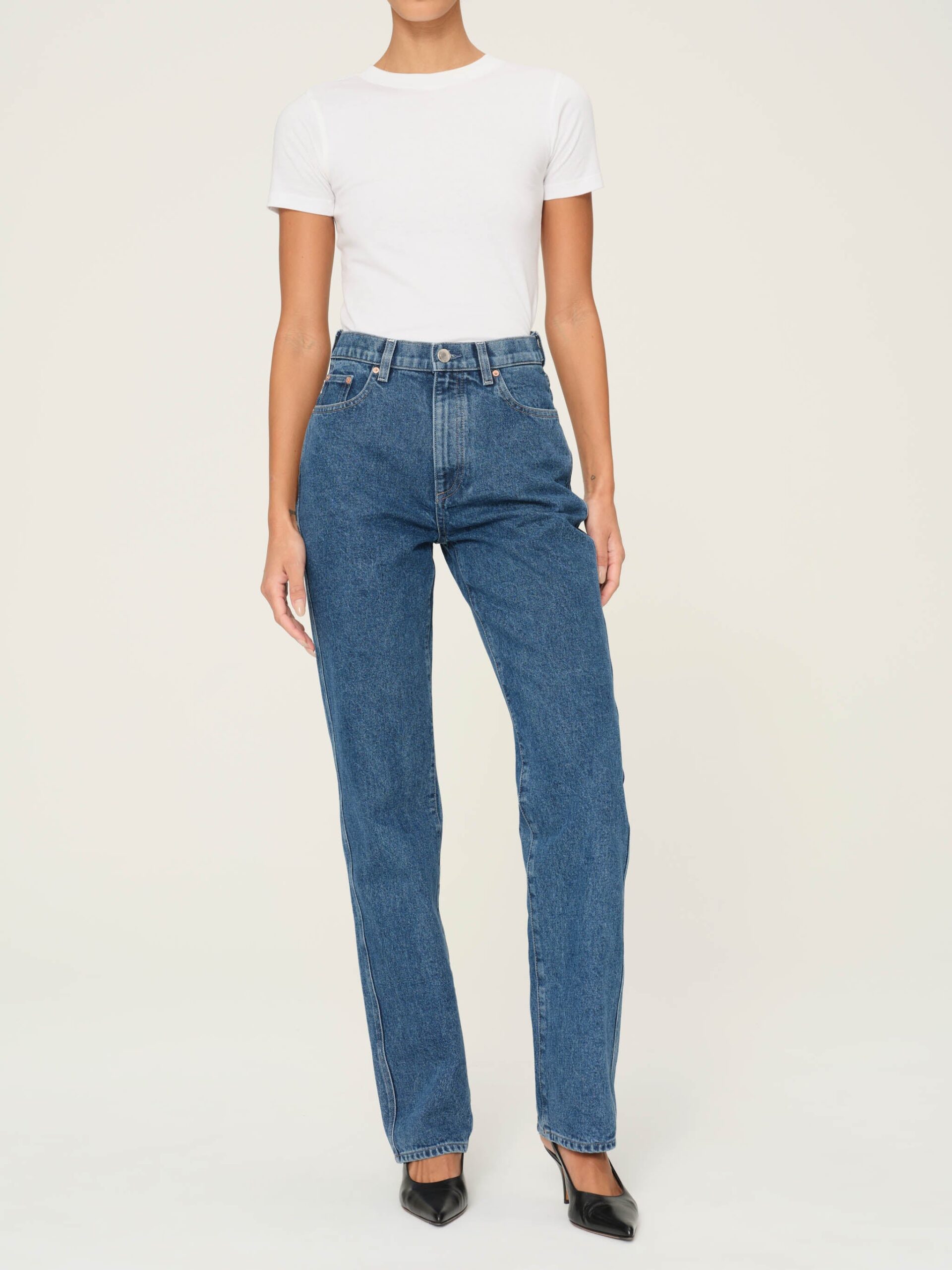 A person wearing a white t-shirt and blue jeans, standing against a plain light-colored background. They are also wearing black pointed-toe shoes.