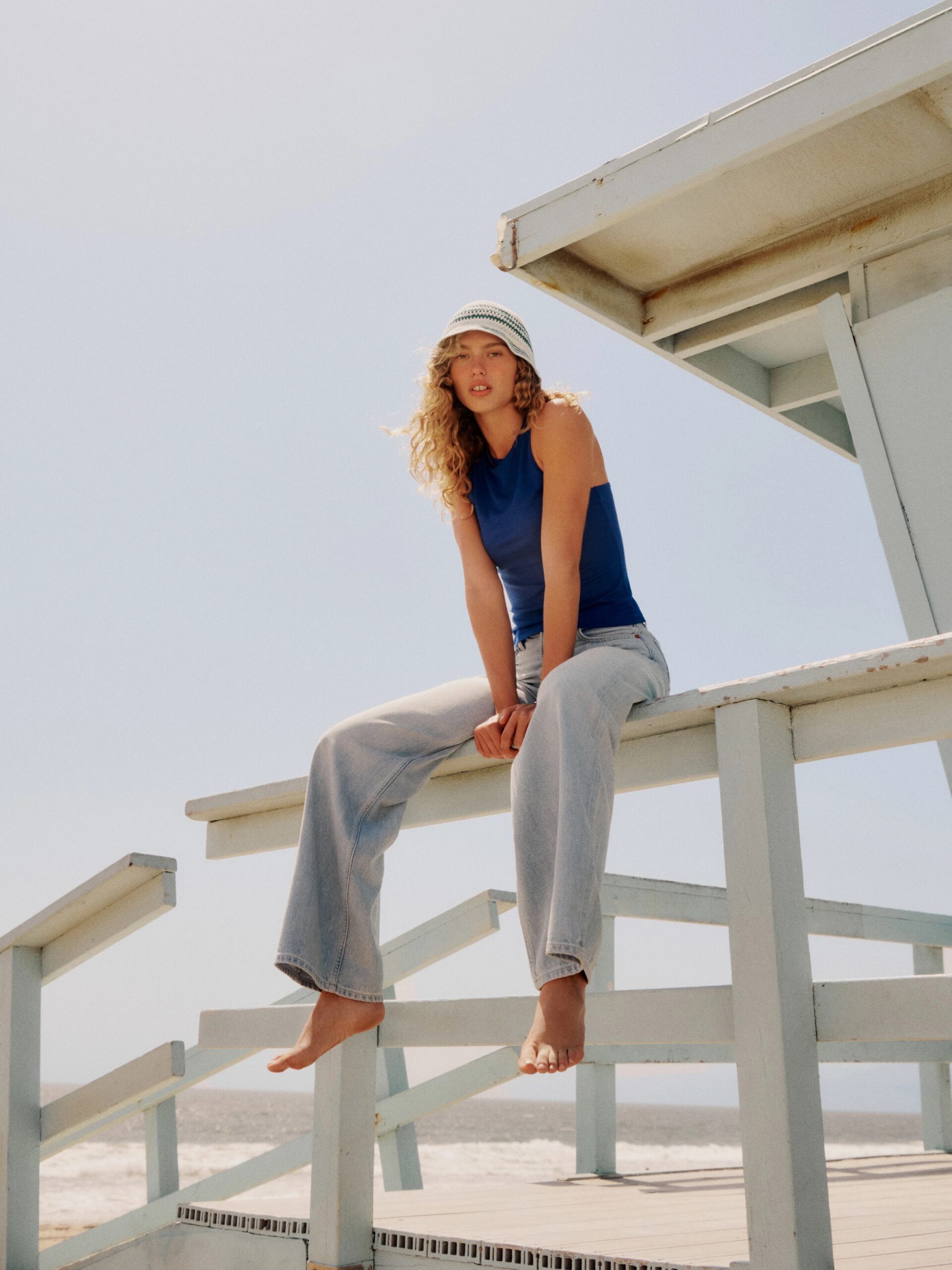 A person wearing a blue tank top, light blue jeans, and a striped bucket hat sits on a lifeguard stand near a beach.